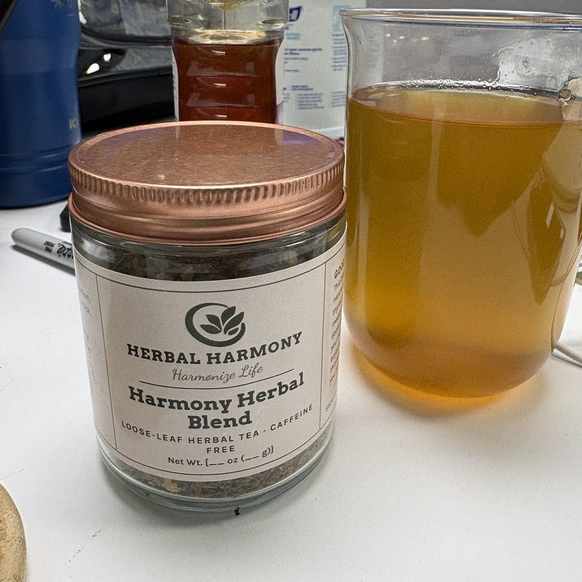 Front view of a Harmony Herbal Blend jar with a rose-gold lid next to a glass mug of golden brewed tea